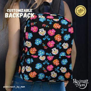 May include: Black backpack with a vibrant sugar skull design. The backpack showcases a repeating pattern of skulls in pink, blue, orange, and yellow, with crossbones and floral details. The text "CUSTOMIZABLE BACKPACK" is displayed on the top left.