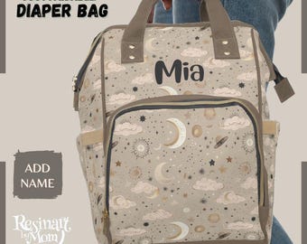 Puede incluir: Un bolso pañalera beige personalizable con un diseño celestial de lunas, estrellas y nubes. El bolso tiene un bolsillo frontal, bolsillos laterales y un asa superior. El nombre "Mia" está impreso en la parte delantera. El bolso es sostenido por una persona.