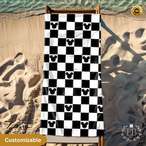 Op de afbeelding: Een zwart-wit geblokte strandlaken met Mickey Mouse silhouetten. De handdoek ligt op een houten strandstoel op een zandstrand. Het woord "Customizable" staat linksonder afgedrukt.