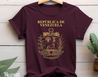 May include: A maroon t-shirt featuring the words "REPÚBLICA DE VENEZUELA" in gold, above a gold emblem. The shirt hangs on a wooden hanger against a white wood-panelled wall. The text "MORE COLORS AVAILABLES" is in the top left corner.