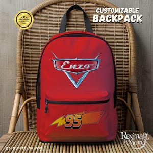 Puede incluir: Mochila roja con cremallera y correas negras, con el texto "Enzo" en un emblema plateado y el n&uacute;mero "95" con un dise&ntilde;o de rayo. La mochila est&aacute; etiquetada como "Mochila personalizable" y "Calidad Premium".