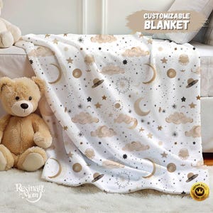 May include: A white blanket with a celestial pattern featuring moons, stars, clouds, and UFOs in gold and black. The blanket has the text "CUSTOMIZABLE BLANKET" and is next to a teddy bear.