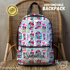 May include: A white customizable backpack with a pink and blue Minnie Mouse print. The backpack has a black zipper and a black strap. The text "create" and "dream" is repeated on the backpack. The name "Samantha" is written in pink glitter on the front of the backpack.