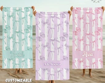 May include: Three rectangular beach towels featuring vertical stripes and a repeating bow pattern. The left towel is light blue with the name Hannah, the centre is light purple with Melissa, and the right is light pink with the name Ava.