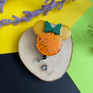 May include: A yellow and orange waffle Mickey Mouse head with a green glitter bow, attached to a clear plastic retractable badge holder with a black and silver clip.