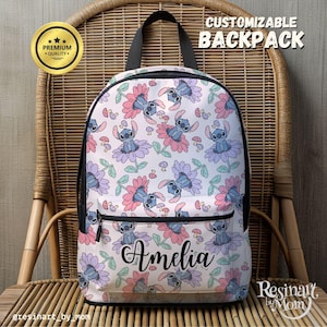 May include: A white backpack with a floral pattern featuring blue Stitch characters from the Disney movie Lilo & Stitch. The backpack has a black zipper and a black adjustable strap. The name "Amelia" is written in black cursive on the front of the backpack.