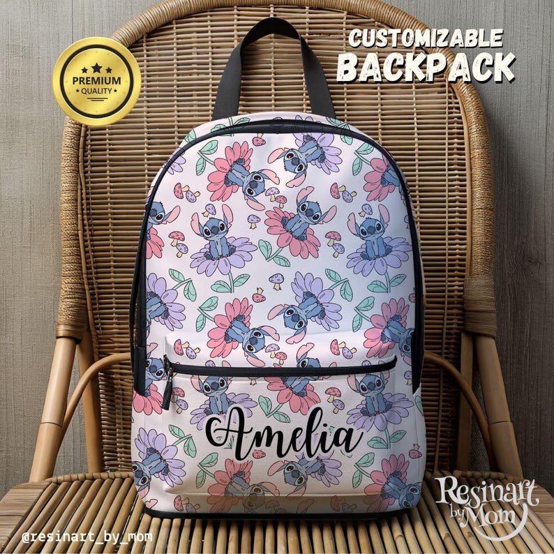 May include: A white backpack with a floral pattern featuring blue Stitch characters from the Disney movie Lilo & Stitch. The backpack has a black zipper and a black adjustable strap. The name "Amelia" is written in black cursive on the front of the backpack.
