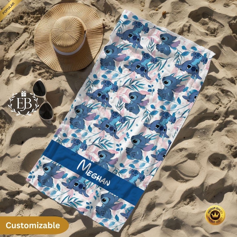May include: A beach towel featuring a pattern of blue cartoon characters and pink and blue foliage on a white background. The towel has a blue stripe with the name "Meghan" in white. A straw hat and sunglasses are next to the towel.