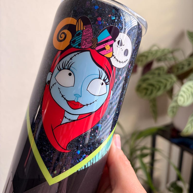 Puede incluir: Un vaso negro con acabado brillante, con un gr&aacute;fico colorido de Sally de The Nightmare Before Christmas. El dise&ntilde;o incluye la cara de Sally, cabello rojo y una diadema con varios patrones. El vaso es sostenido por una mano.