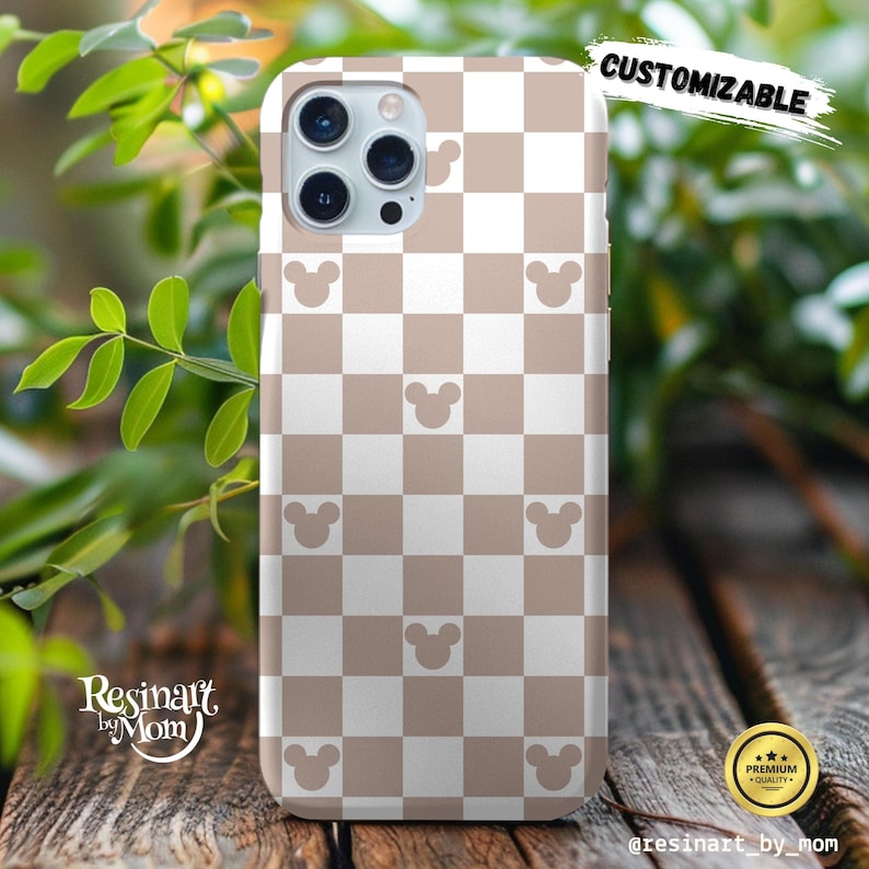 May include: A phone case with a tan and white checkered pattern, featuring Mickey Mouse silhouettes. The case is labeled "CUSTOMIZABLE" and has the text "Resinart by Mom" on it. The case is on a wooden surface with green foliage in the background.