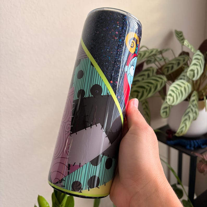 Puede incluir: Un vaso con un dise&ntilde;o inspirado en Sally de Pesadilla antes de Navidad. El vaso tiene una parte superior de purpurina azul oscuro y un dise&ntilde;o de patchwork en negro, morado y verde azulado. Una raya amarilla acent&uacute;a el dise&ntilde;o.