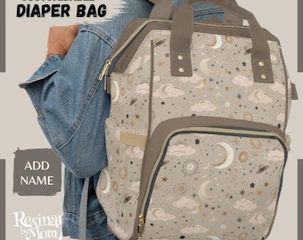 Puede incluir: Una bolsa de pañales beige con un estampado celestial de lunas, estrellas y nubes. La bolsa tiene detalles marrones, un bolsillo frontal y un asa superior. El texto "CUSTOMIZABLE DIAPER BAG" está impreso en la parte superior.