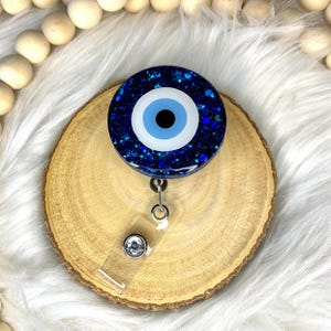 May include: A circular badge reel featuring a dark blue resin base filled with shimmering glitter. The center displays a classic evil eye motif with a black pupil, a light blue iris, and a white outer ring. A clear plastic strap with a metal snap.