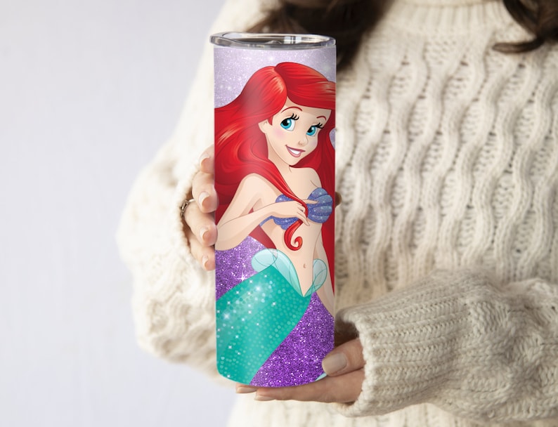 May include: A stainless steel tumbler with a purple glitter background and a colorful image of Ariel, the Little Mermaid, with a seashell bra and a green tail.