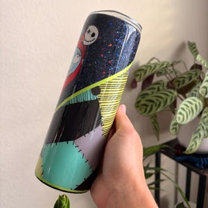 Puede incluir: Un vaso de acero inoxidable con un dise&ntilde;o inspirado en "Pesadilla antes de Navidad". El vaso tiene un fondo negro brillante con im&aacute;genes de Jack Skellington y Sally. El dise&ntilde;o incluye patrones de patchwork y rayas.