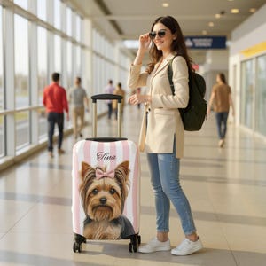 Puede incluir: Una maleta con ruedas que presenta un patrón de rayas verticales en rosa claro y blanco con el gráfico de un Yorkshire Terrier con un lazo rosa. El nombre Tina está impreso en cursiva negra sobre el perro. La maleta tiene un asa retráctil.