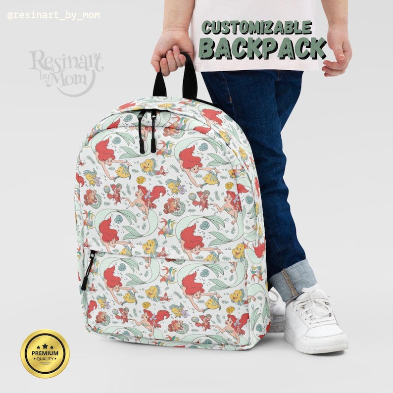 Puede incluir: Una mochila blanca con un patr&oacute;n colorido de sirenas, peces y conchas. La mochila tiene correas negras y una cremallera negra. El texto "MOCHILA PERSONALIZABLE" est&aacute; impreso en la parte posterior de la mochila.