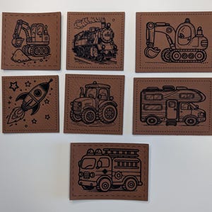 Könnte beinhalten: Sieben braune Lederaufnäher mit schwarzen Strichzeichnungen von Fahrzeugen und einer Rakete. Zu den Fahrzeugen gehören ein Bagger, ein Zug, ein Traktor, ein Wohnmobil und ein Feuerwehrauto. Die Rakete ist von Sternen umgeben.