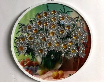 May include: A white framed round wall art piece with a colorful image of a bouquet of daisies with yellow centers, green leaves, and a vase. The daisies are made of rhinestones and beads. The image also includes a bowl of strawberries and two peaches.