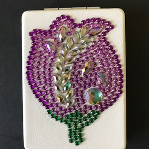 May include: A white compact mirror with a rose design made of purple, green, and iridescent rhinestones.