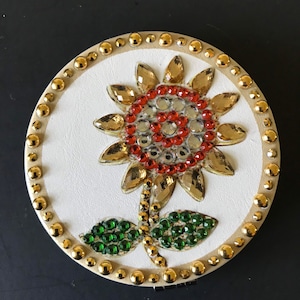 May include: A round, white, decorative trinket box with a gold trim. The lid features a sunflower design made of rhinestones in shades of gold, red, and green.