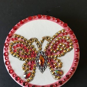 May include: A round, white compact with a pink border and a butterfly design made of red, gold, and clear rhinestones.