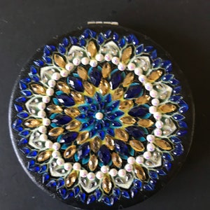 May include: A round, black compact mirror with a blue, gold, and white jeweled design. The mirror has a circular, jeweled pattern with a central white pearl.