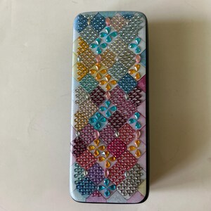 May include: A small, rectangular box with a colorful, geometric pattern. The pattern is made up of small, iridescent beads in shades of blue, green, pink, and yellow. The box has a black border.