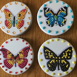 May include: Four round, white compact mirrors with butterfly designs. Each mirror has a different colored butterfly with rhinestones. The butterflies are orange, blue, red, and black.