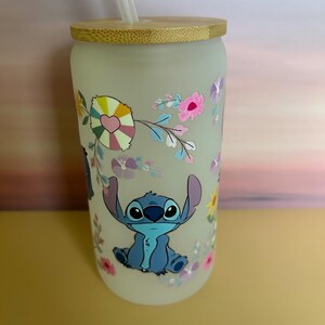 Op de afbeelding: Matglazen beker met een bamboe deksel en rietje. De beker heeft een kleurrijk bloemmotief en een stripfiguur. Het personage is blauw met grote ogen en oren. De bloemen zijn roze, geel en paars.