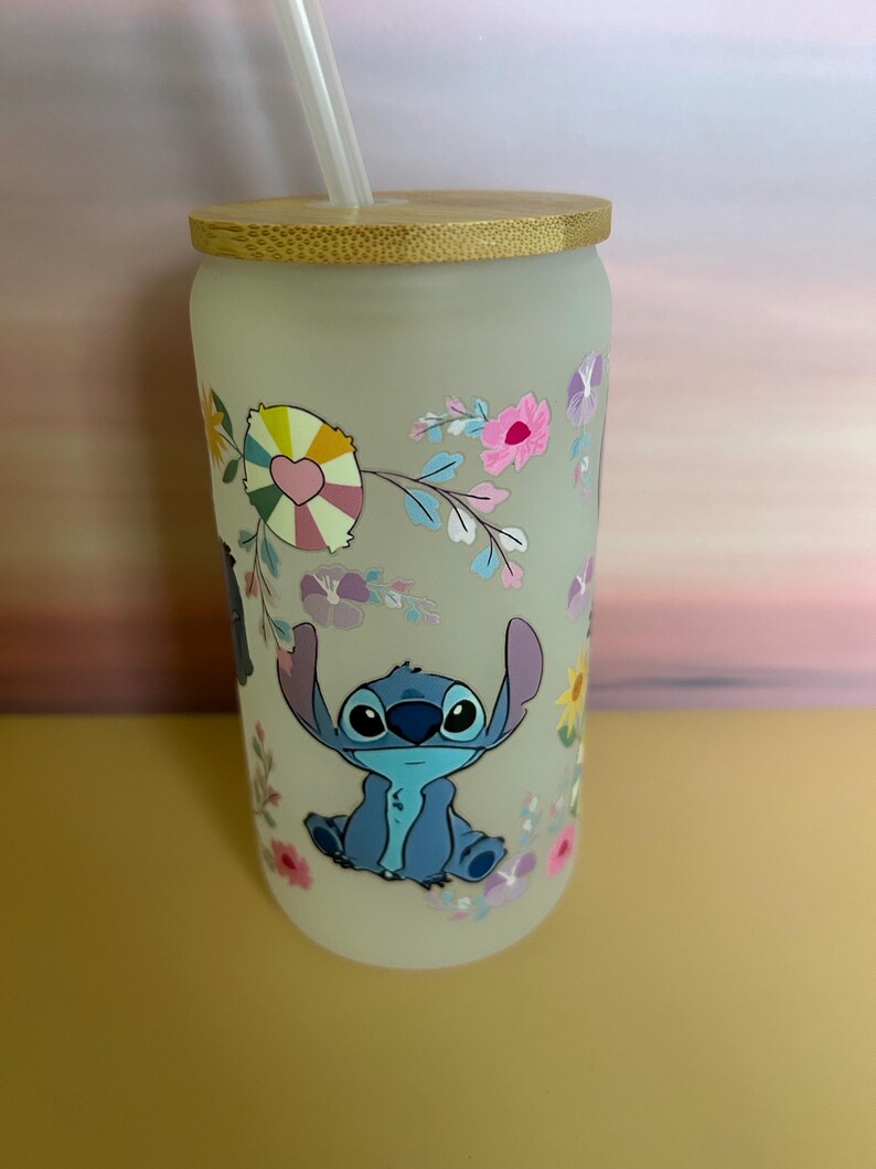 Op de afbeelding: Matglazen beker met een bamboe deksel en rietje. De beker heeft een kleurrijk bloemmotief en een stripfiguur. Het personage is blauw met grote ogen en oren. De bloemen zijn roze, geel en paars.
