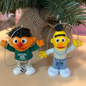 May include: Two Sesame Street character ornaments. One is orange with black hair and a green shirt that says "Sesame 123 Street". The other is yellow with a black mohawk and a white and gray outfit. Both have silver hanging loops.