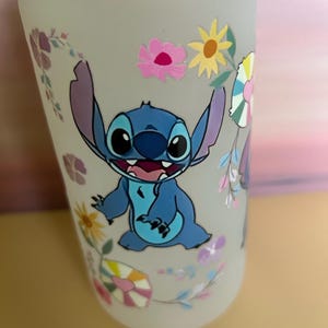 Op de afbeelding: Een frosted glazen waterfles met een blauw stripfiguur en bloemmotieven. Het figuur heeft grote oren en een brede glimlach. De bloemen zijn roze, geel en paars.