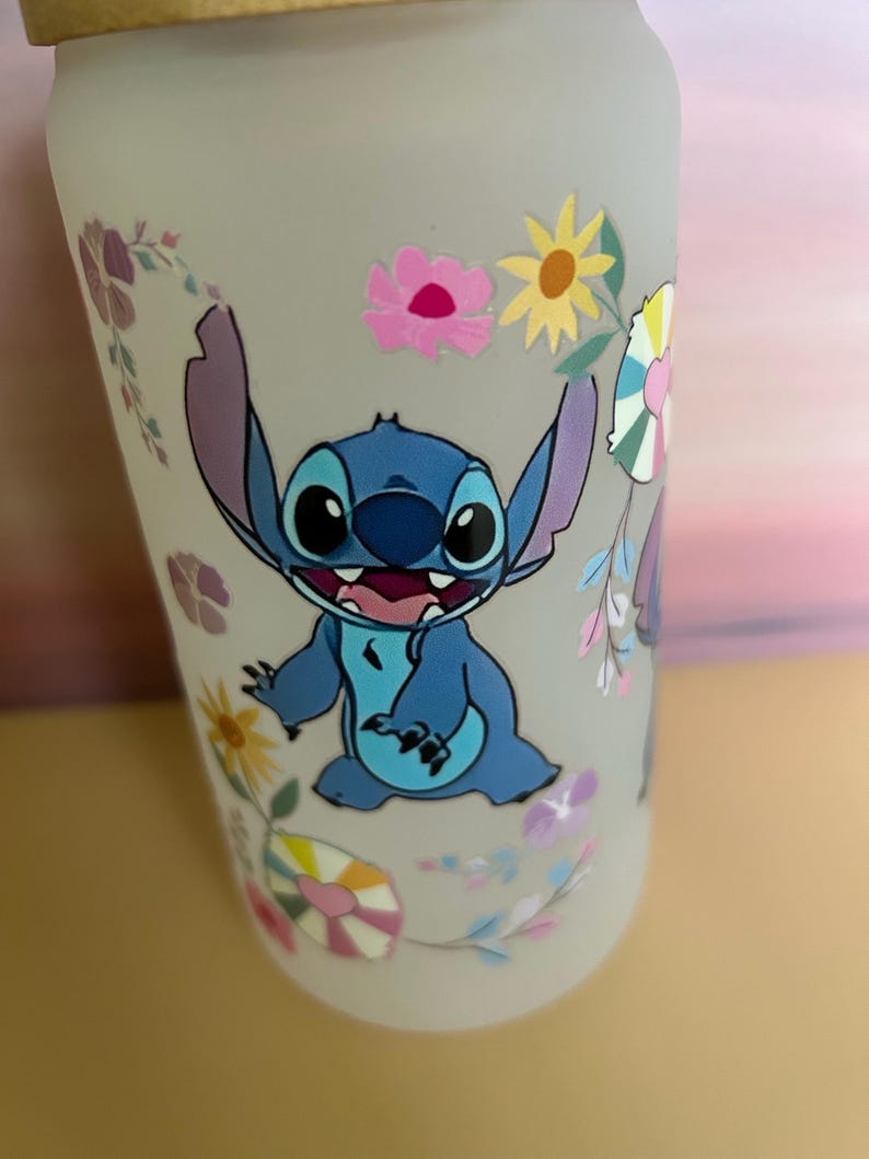 Op de afbeelding: Een frosted glazen waterfles met een blauw stripfiguur en bloemmotieven. Het figuur heeft grote oren en een brede glimlach. De bloemen zijn roze, geel en paars.