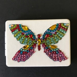 May include: A white compact mirror with a colorful butterfly design made of rhinestones. The butterfly has green, blue, red, yellow, and pink rhinestones.