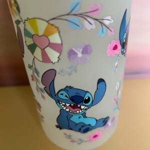 Op de afbeelding: Een frosted beker met een lachend blauw stripfiguur met grote oren en een brede grijns. De beker is versierd met kleurrijke bloemen- en hartvormige ontwerpen. Het algehele ontwerp is grillig en speels.