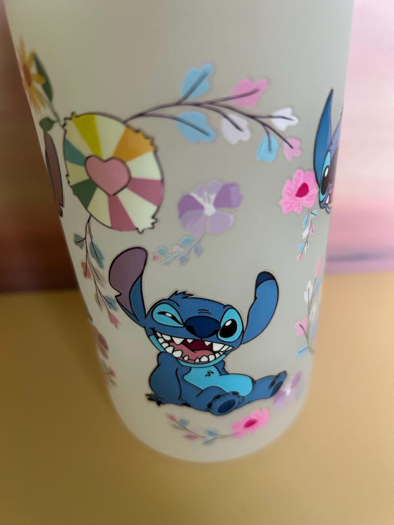 Op de afbeelding: Een frosted beker met een lachend blauw stripfiguur met grote oren en een brede grijns. De beker is versierd met kleurrijke bloemen- en hartvormige ontwerpen. Het algehele ontwerp is grillig en speels.