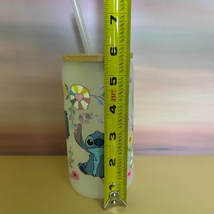 Op de afbeelding: Een heldere glazen beker met een bamboe deksel en rietje, met een cartoon karakter ontwerp. De beker is ongeveer 15 cm hoog. Het ontwerp bevat Stitch, bloemen en kleurrijke accenten. Een perfect cadeau voor fans.