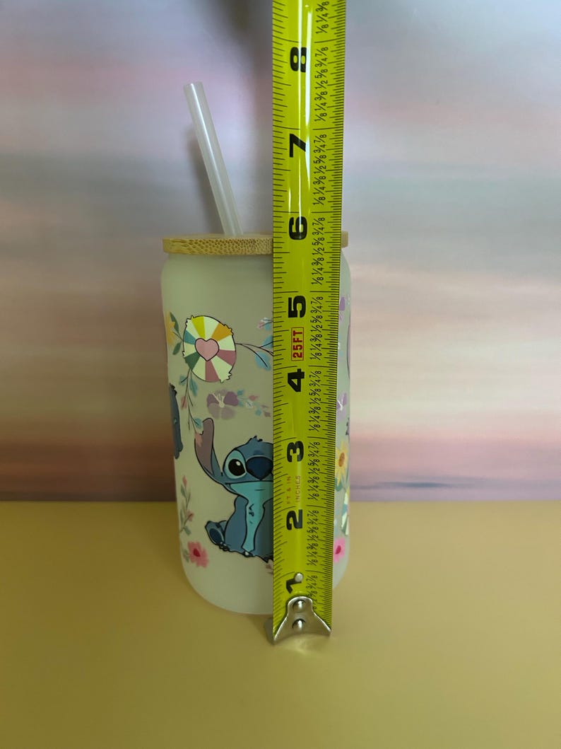 Op de afbeelding: Een heldere glazen beker met een bamboe deksel en rietje, met een cartoon karakter ontwerp. De beker is ongeveer 15 cm hoog. Het ontwerp bevat Stitch, bloemen en kleurrijke accenten. Een perfect cadeau voor fans.