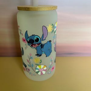 Op de afbeelding: Matte glazen beker met een blauw stripfiguur, grote oren en een vrolijke uitdrukking, omringd door bloemen- en hartmotieven. De beker heeft een bamboe deksel en een doorzichtig rietje. Een leuk en kleurrijk drinkartikel.