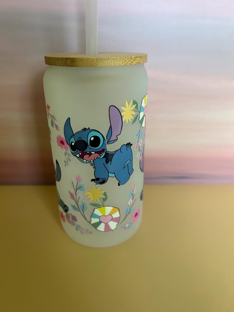 Op de afbeelding: Matte glazen beker met een blauw stripfiguur, grote oren en een vrolijke uitdrukking, omringd door bloemen- en hartmotieven. De beker heeft een bamboe deksel en een doorzichtig rietje. Een leuk en kleurrijk drinkartikel.