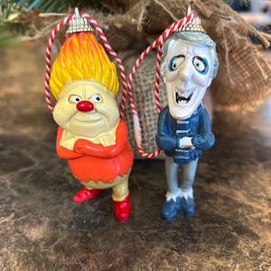 May include: Two Christmas tree ornaments. One is a cartoon character with orange hair, a red nose, and a red outfit. The other is a cartoon character with blue clothing and white hair. Both ornaments have red and white string hangers.