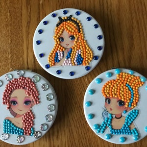 May include: Three round, white compact mirrors with a different cartoon princess on each. The princess on the left has red hair and a blue dress. The princess in the middle has blonde hair and a blue and pink dress. The princess on the right has blonde hair and a blue dress.