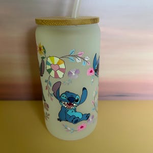 Op de afbeelding: Matglazen beker met een bamboe deksel en rietje. De beker heeft een kleurrijk bloemenontwerp en een stripfiguur. Het personage is blauw met grote oren en een brede grijns. De beker is ontworpen voor koude dranken.