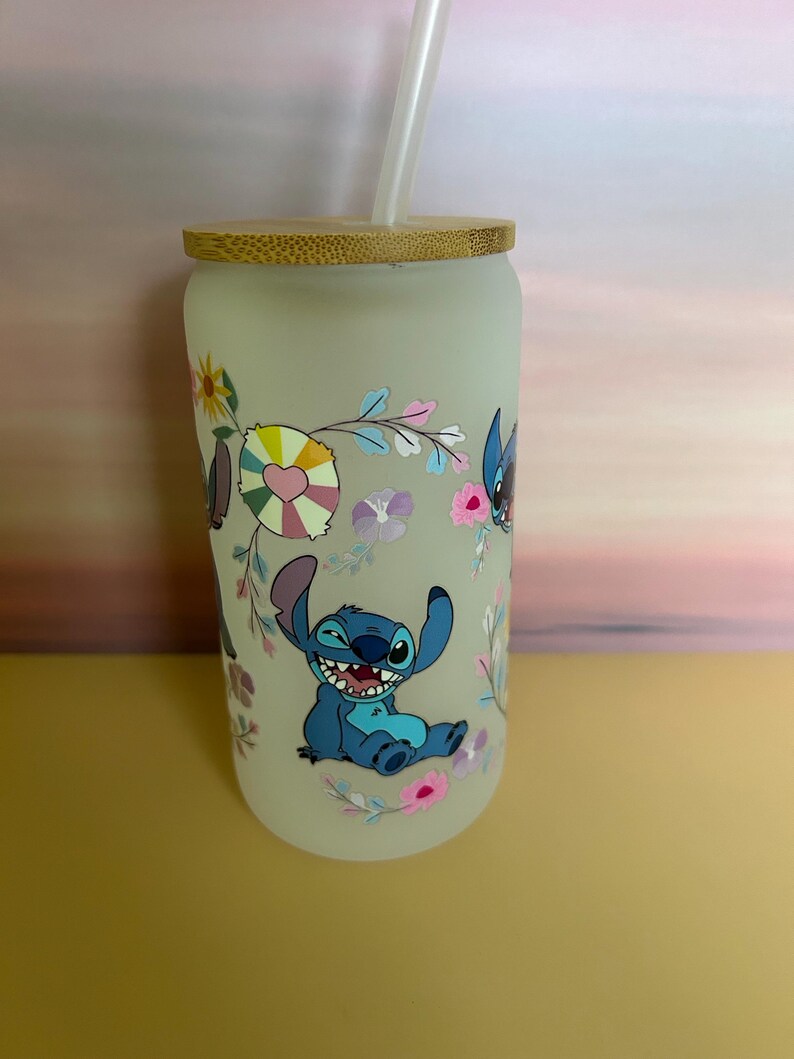 Op de afbeelding: Matglazen beker met een bamboe deksel en rietje. De beker heeft een kleurrijk bloemenontwerp en een stripfiguur. Het personage is blauw met grote oren en een brede grijns. De beker is ontworpen voor koude dranken.