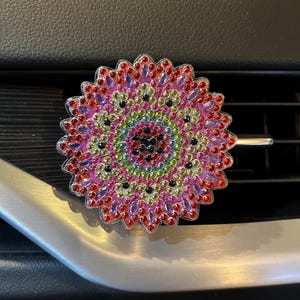 May include: A colorful car air vent clip featuring a circular, mandala-like design. The clip is adorned with numerous small, sparkling rhinestones in shades of red, pink, purple, green, and black. The clip is silver and is attached to a car vent.
