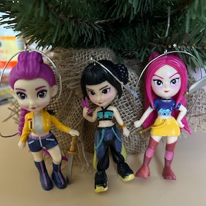May include: Three Christmas tree ornaments featuring stylized female figures with vibrant hair and outfits. The figures have purple, black, and pink hair, and wear colorful clothing. Each ornament has a silver loop for hanging.