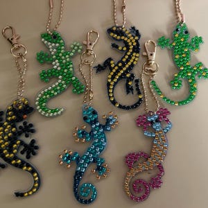 May include: A collection of lizard-shaped keychains in various colors, including green, blue, yellow, and pink. Each keychain is adorned with sparkling embellishments and attached to a gold-colored clasp and chain. The keychains are designed with intricate details.