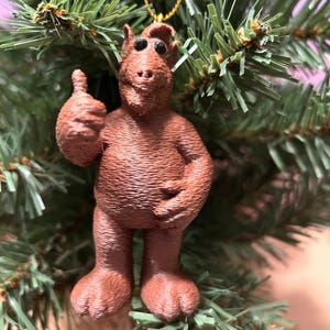 May include: A brown, 3D-printed ornament of a cartoon character giving a thumbs-up. The ornament has a textured surface and is hanging from a gold string. The ornament is displayed on a green Christmas tree.
