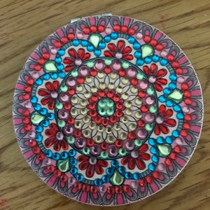 May include: A round, pink compact mirror with a colorful mandala design. The design features a variety of colors, including red, blue, green, and yellow. The mirror is decorated with small, round rhinestones.